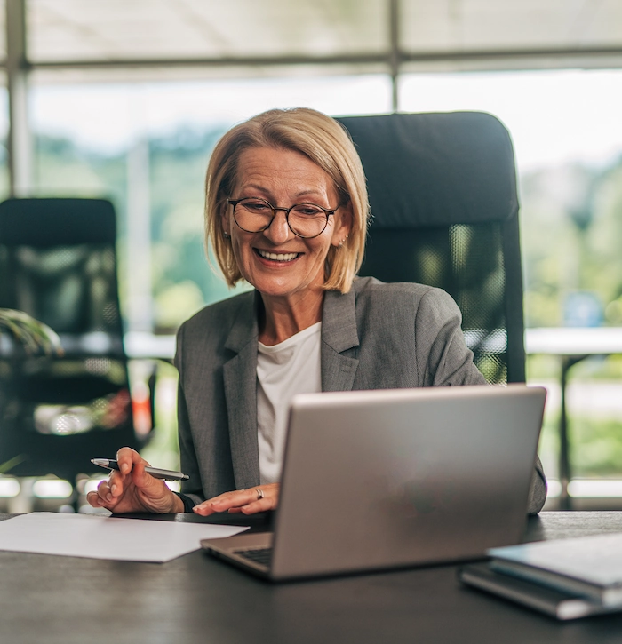 Blonde vrouw met bril werkt glimlachend achter een laptop op kantoor.
