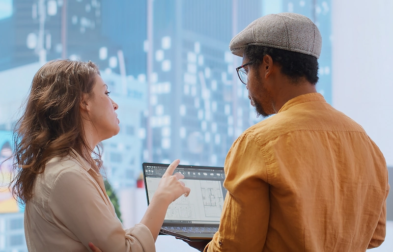 Twee collega's overleggen samen terwijl ze bouwtekeningen op een laptop bekijken.