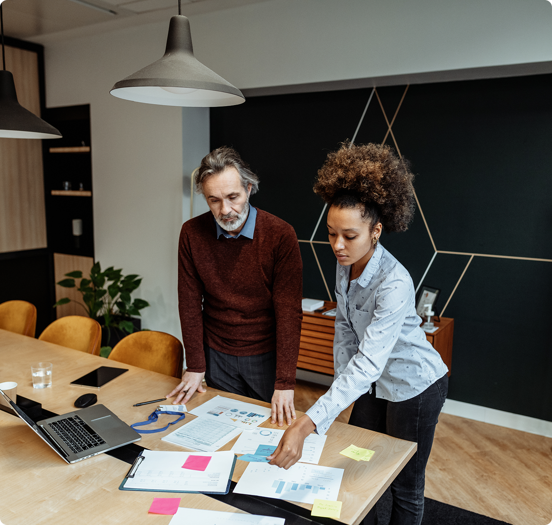 Twee collega's werken samen aan een tafel vol documenten, grafieken en aantekeningen.