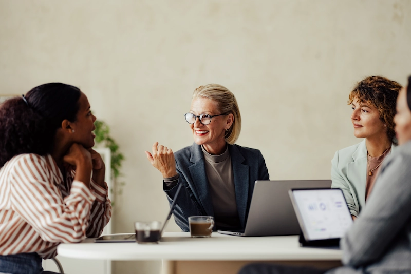 Een groep collega’s in gesprek rond een tafel, met een vrouw in het midden die enthousiast praat.