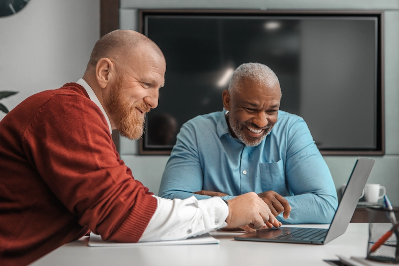Twee collega’s werken samen lachend achter een laptop in een vergaderruimte.