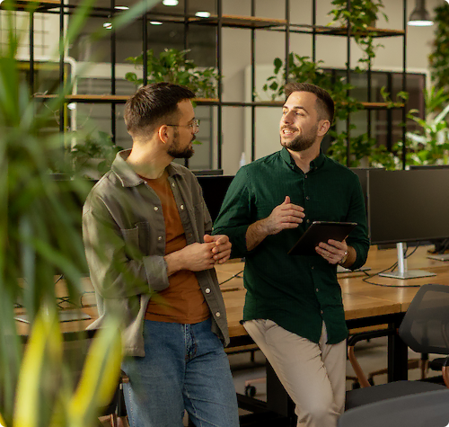 Twee mannelijke collega’s praten met elkaar in een modern kantoor met veel planten.