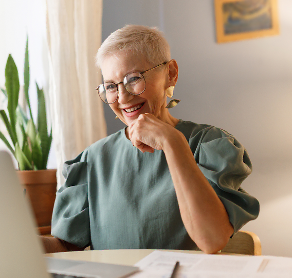 Vrouw met kort wit haar werkt glimlachend achter een laptop aan een bureau.