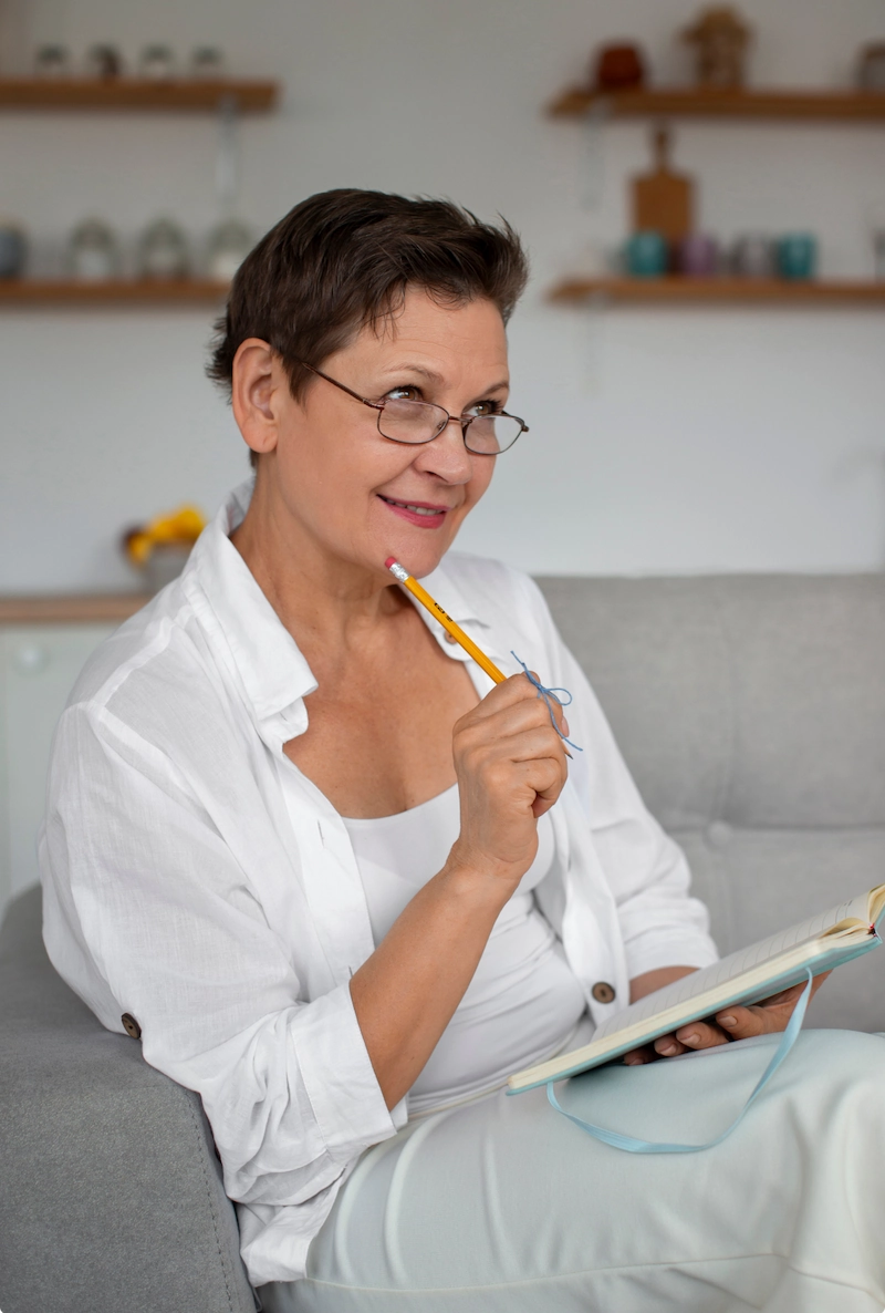 Vrouw met bril denkt na terwijl ze met een potlood in de hand in een notitieboek schrijft.